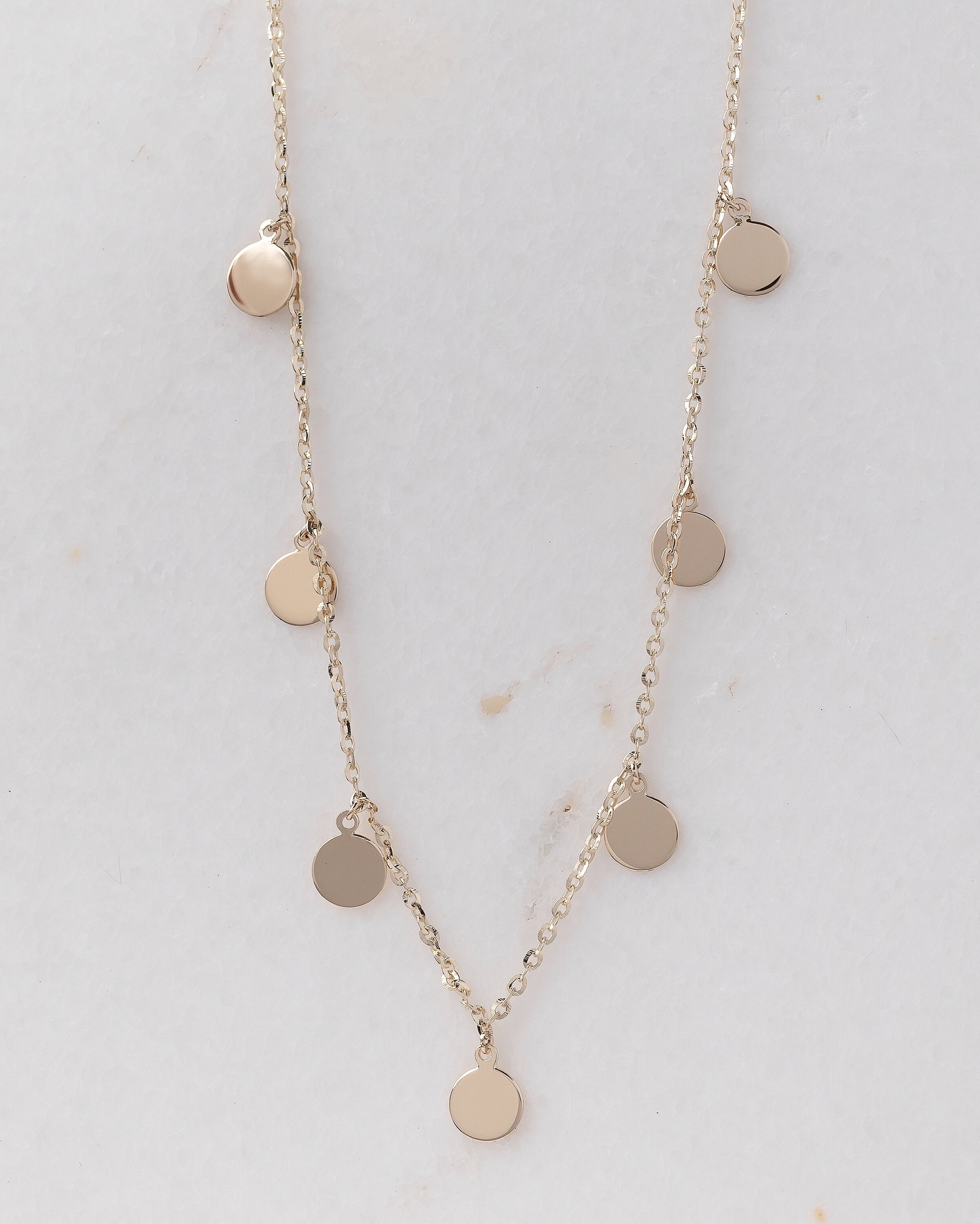 The photo shows a delicate gold necklace featuring multiple small, round, flat coin-shaped pendants evenly spaced along a fine gold chain. 