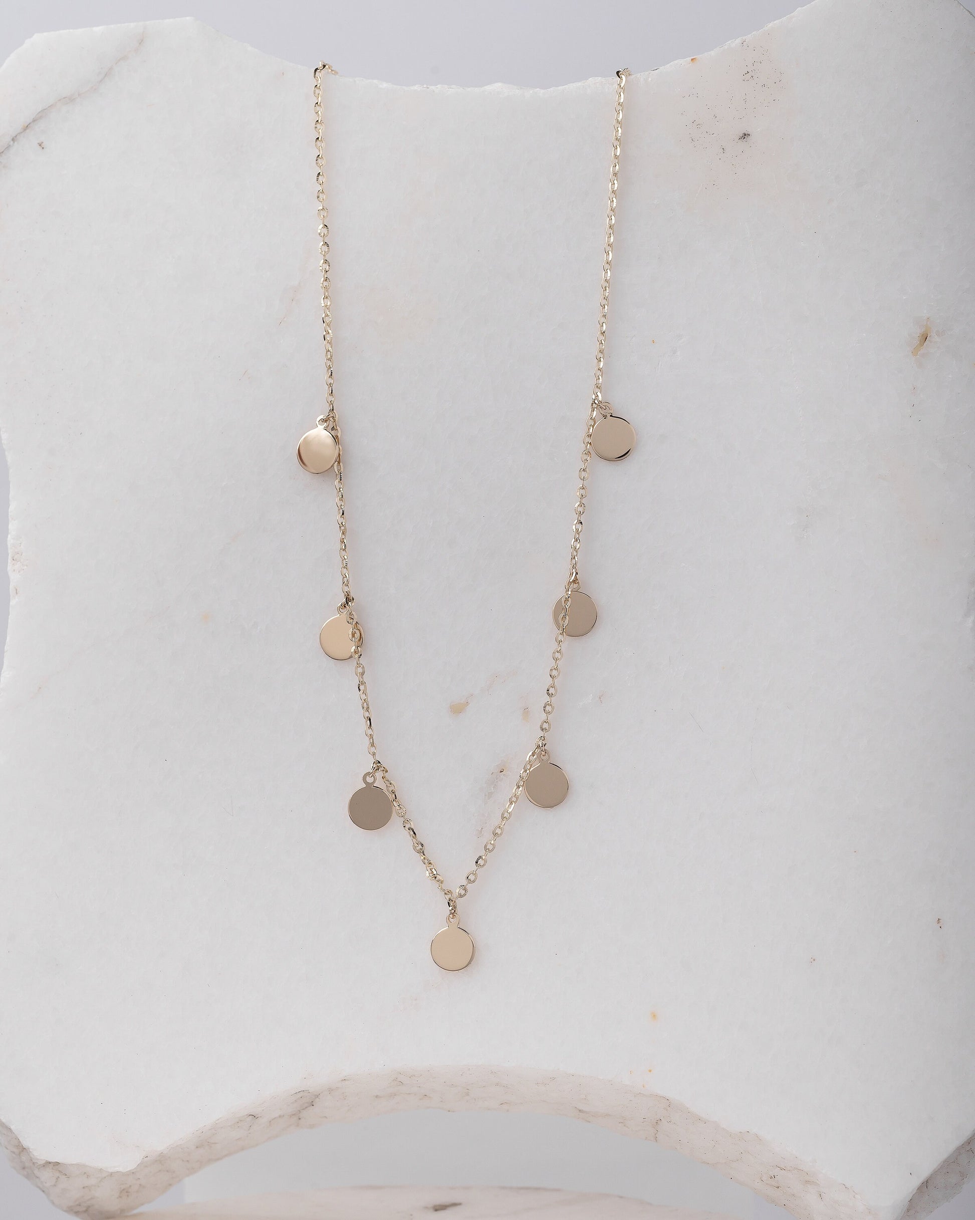 The photo shows a delicate gold necklace featuring multiple small, round, flat coin-shaped pendants evenly spaced along a fine gold chain. 