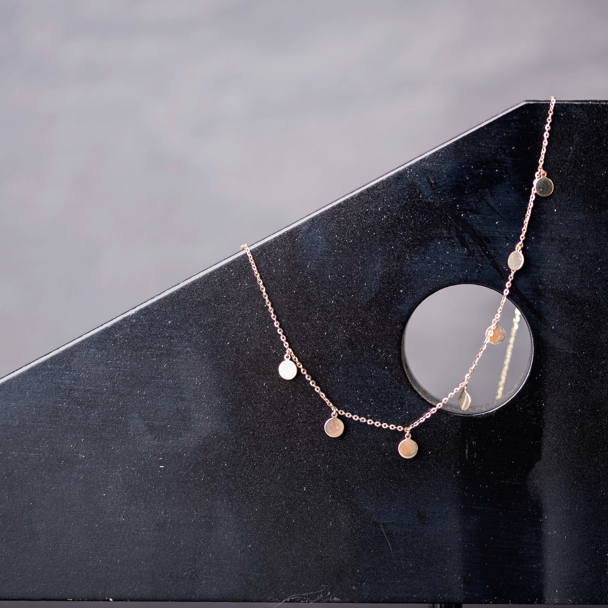 The photo shows a delicate gold necklace featuring multiple small, round, flat coin-shaped pendants evenly spaced along a fine gold chain. 