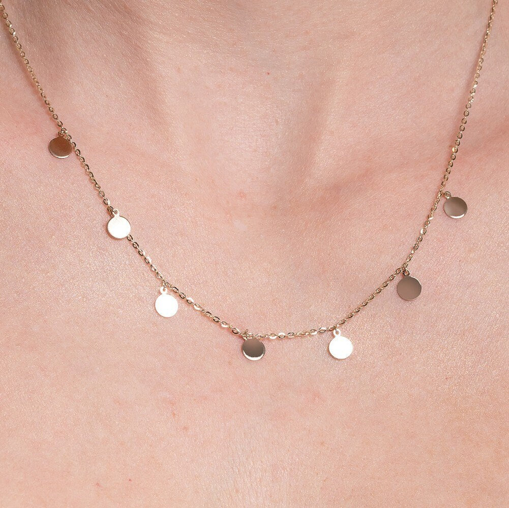 The photo shows a delicate gold necklace featuring multiple small, round, flat coin-shaped pendants evenly spaced along a fine gold chain. 