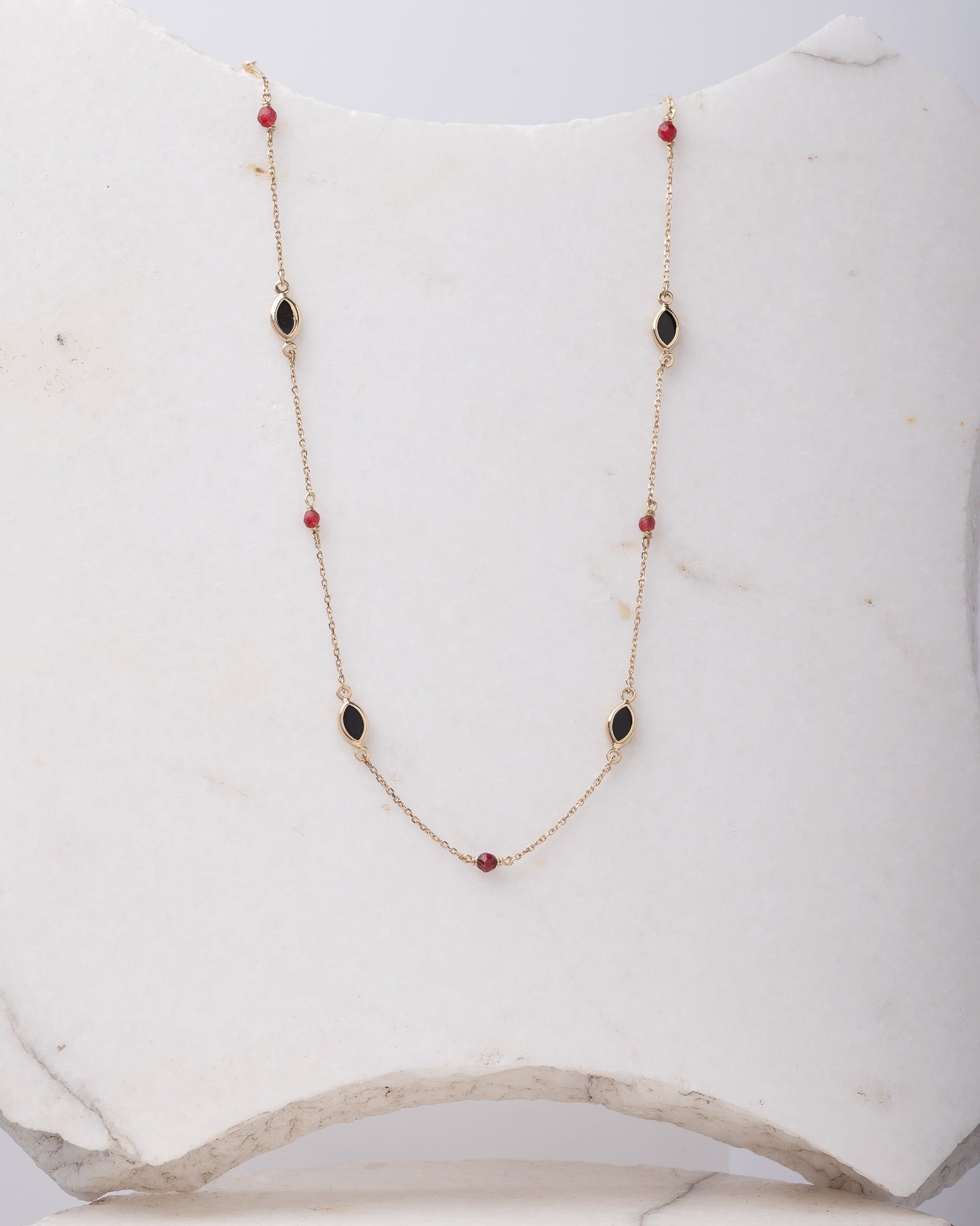 The photo shows a delicate gold necklace featuring small tourmaline gemstones and black marquise-shaped onyx accents, spaced along a fine chain. The contrasting colors create a striking and elegant design