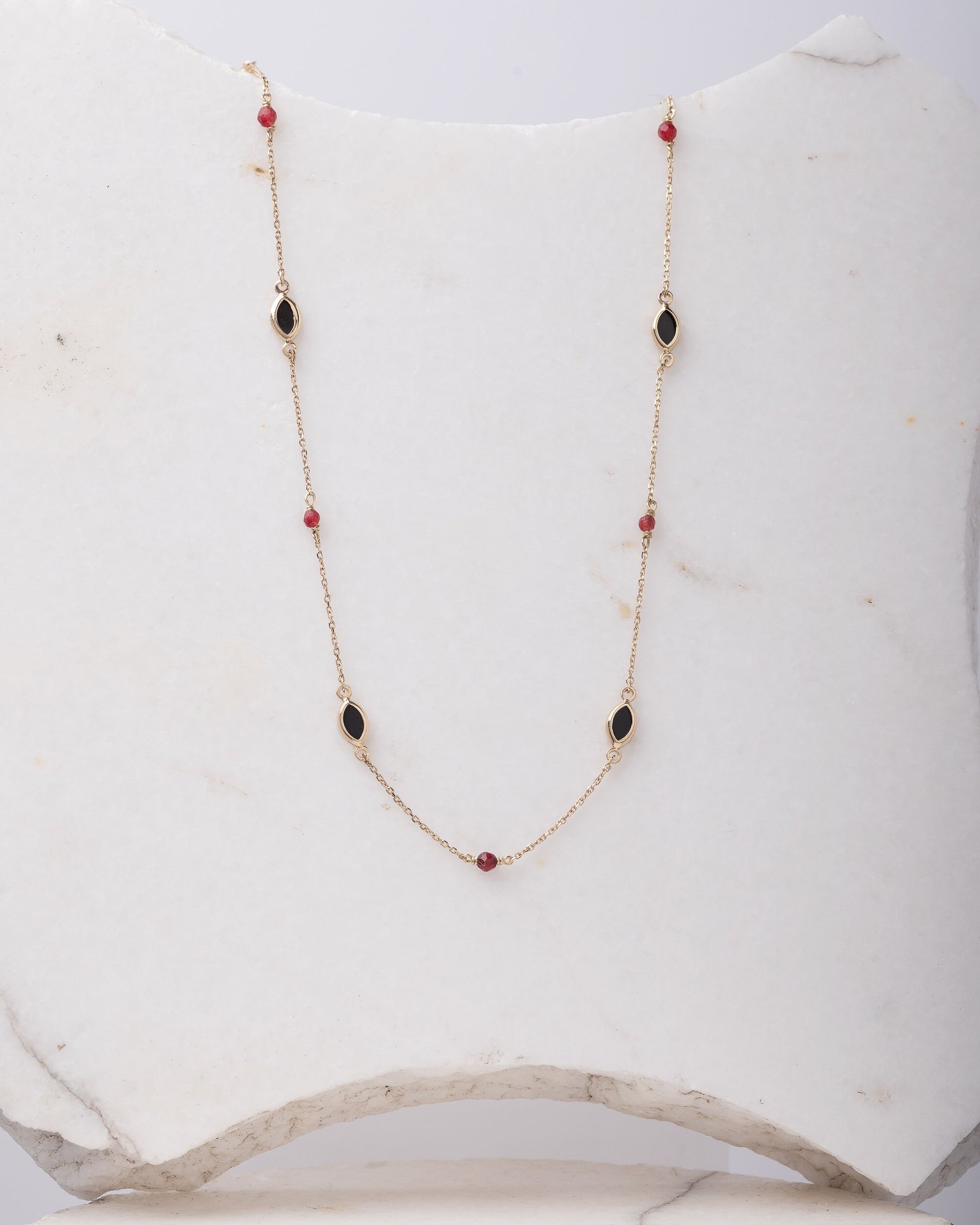 The photo shows a delicate gold necklace featuring small tourmaline gemstones and black marquise-shaped onyx accents, spaced along a fine chain. The contrasting colors create a striking and elegant design