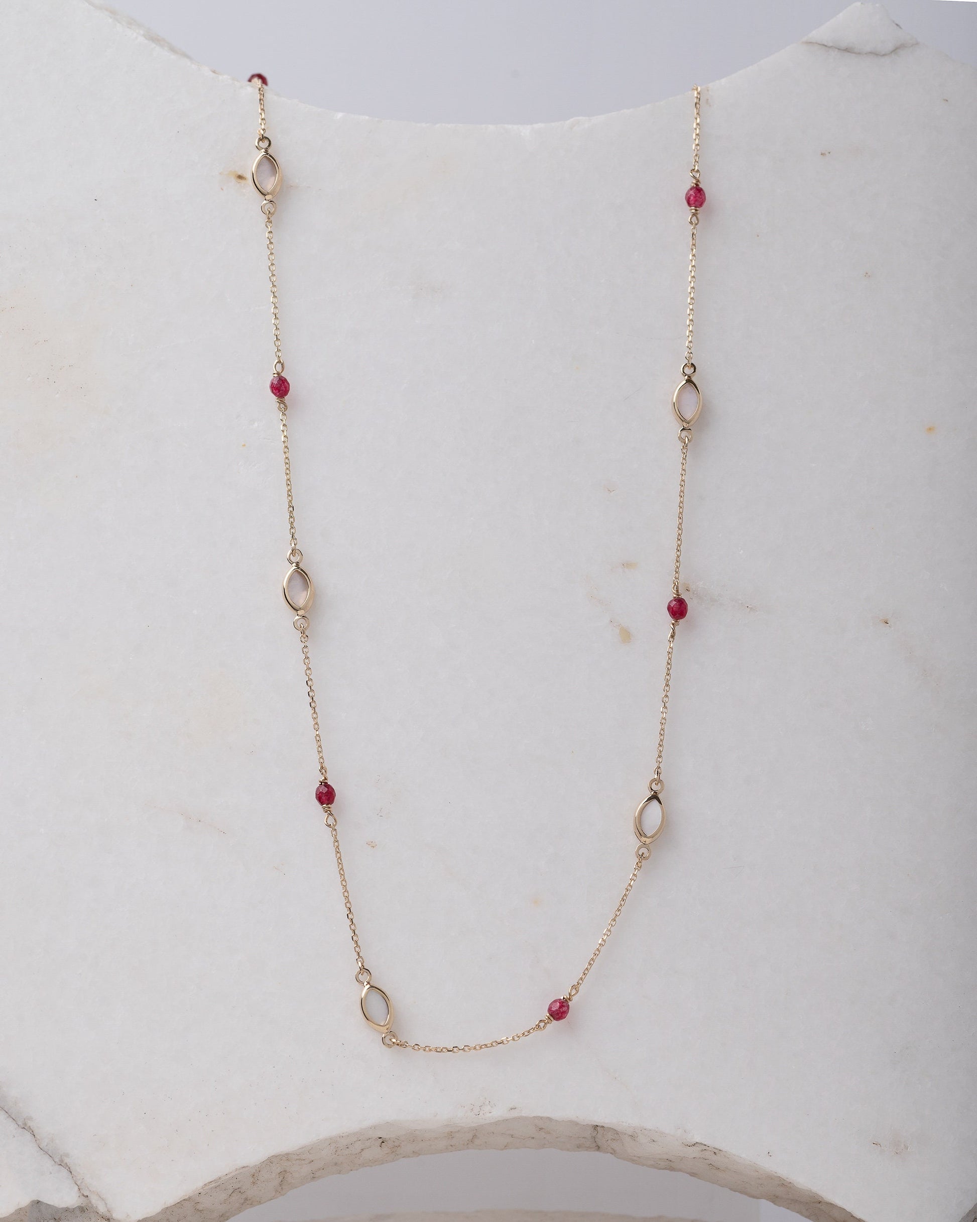 The photo shows a delicate gold necklace featuring small tourmaline gemstones and black marquise-shaped mother of pearl accents, spaced along a fine chain. The contrasting colors create a striking and elegant design